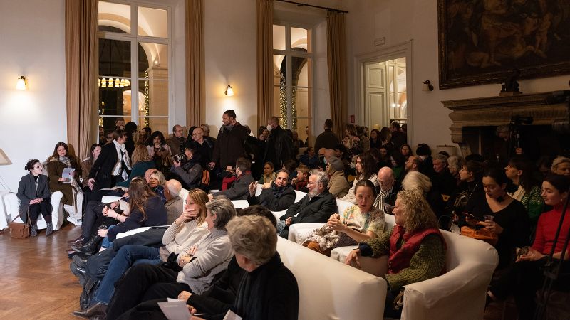 Color photograph of several dozen people seated and standing in a salon, watching a pianist perform