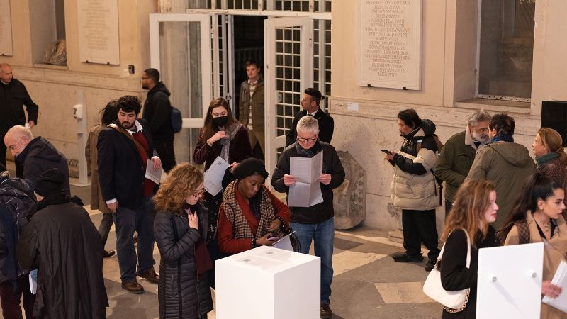 Color photograph of people in the entrance hall to a building, looking at paper handouts taken from a white podium