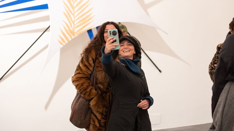 Color photograph of two women taking a selfie in front of an abstract sculpture hanging on a white gallery wall