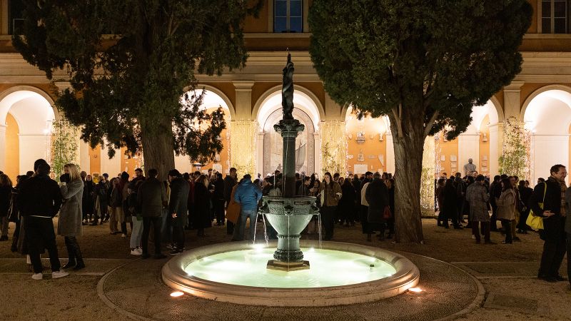 Color photograph of dozens of people socializing around a circular fountain in a courtyard at night