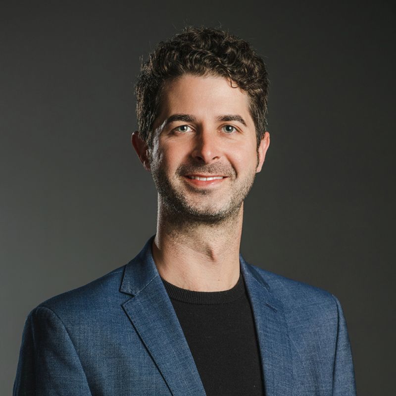 Color photograph of the head and torso of a light skinned man wearing a blue blazer over a black shirt; he smiles and looks at the camera