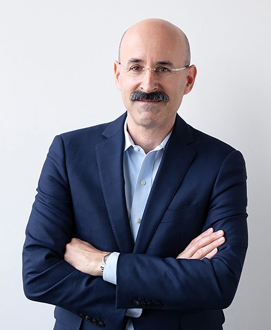 Color photograph of a light skinned man with a bushy mustache seen from the waist up; he wears a dark blazer and glasses and crosses his arms as he smiles at the camera