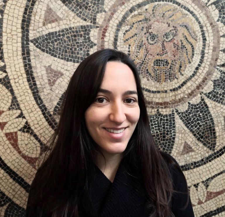 Color photograph of the head and shoulders of Adriana Maria Vazquez standing in front of an ancient Roman mosaic