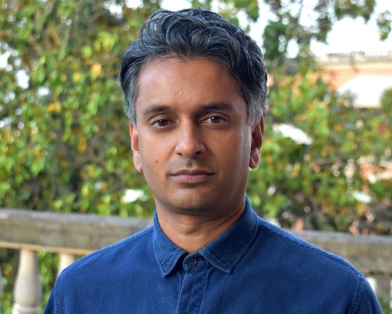 Color photograph of an olive skinned man wearing a blue collared shirt, sitting in a garden, and looking at the camera with a serious expression