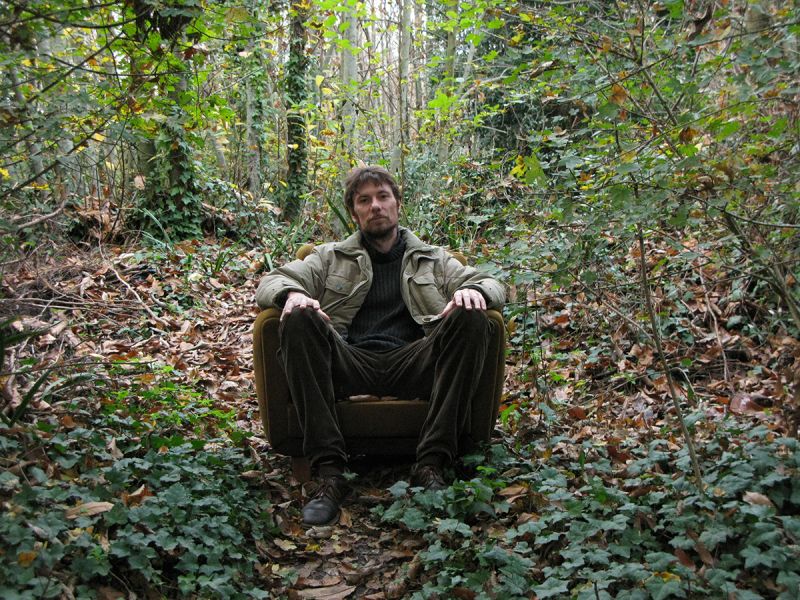 Color photograph of a light skinned man with brown hair and beard sitting in an easy chair in the middle of a forest and looking directly at the camera