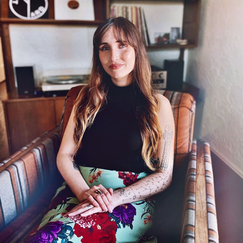 A color photograph of a light skinned woman with auburn hair sitting in a colorful striped chair; she wears a black top, a colorful print skirt, and a sleeveless black top that shows arm tattoos; behind her is a wooden shelf containing a turntable and records