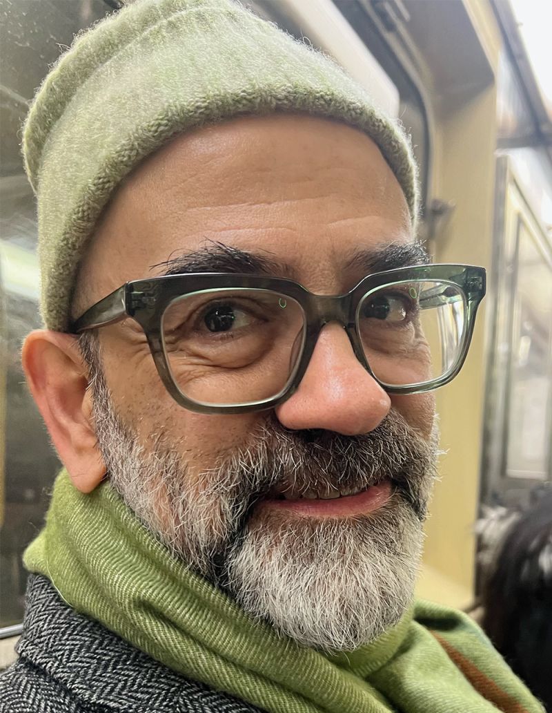 Color photo of the head and shoulders of a light skinned man smiling at the camera; he wears glasses, a cap, and a beard