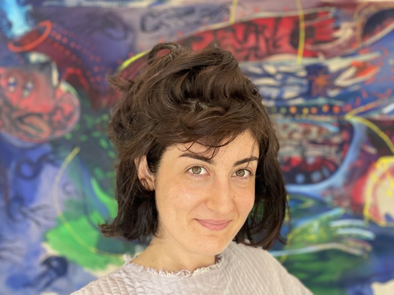 Color photograph of the head of a light skinned woman with brown hair; she is positioned in front of a colorful abstract painting and smiles at the camera