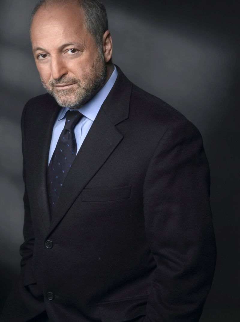 Color photograph of the head and torso of André Aciman, a man wearing a dark suit and tie and standing in front of a dark, shadowy backgroud, looking straight at the camera