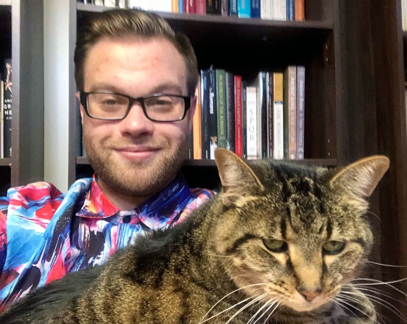 Color photograph of a light skinned man wearing glasses and a multicolored shirt sitting in front of a full bookshelf and smiling at the camera; in his lap he holds a furry cat, which takes up most of the foreground in the picture