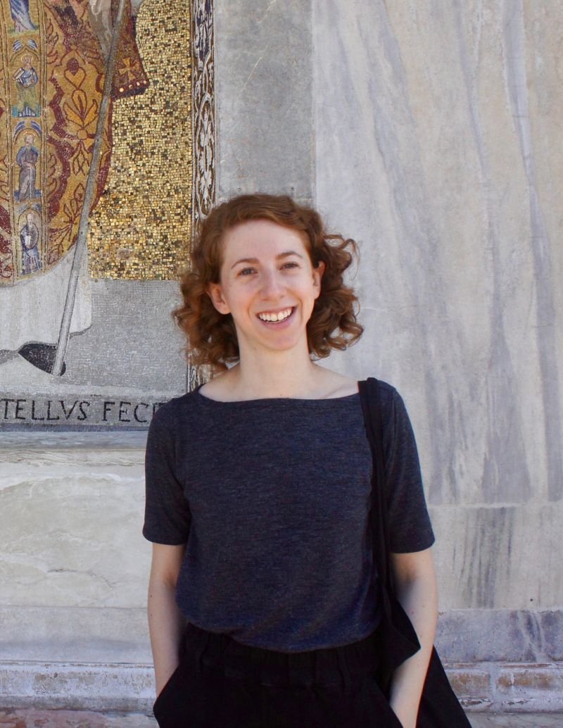 Color portrait of Anna Dumont from the hips up, wearing a gray top, facing forward, and standing in front of a marble wall with a mosaic over her right shoulder