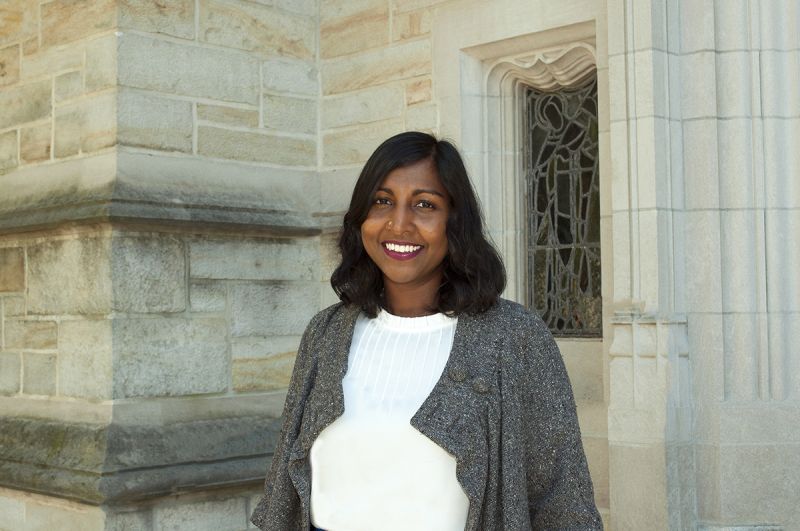 Color photograph of a brown-skinned, dark-haired woman wearing a white top and gray sweater; she stands outdoors in front of the stone wall and glass window of a building