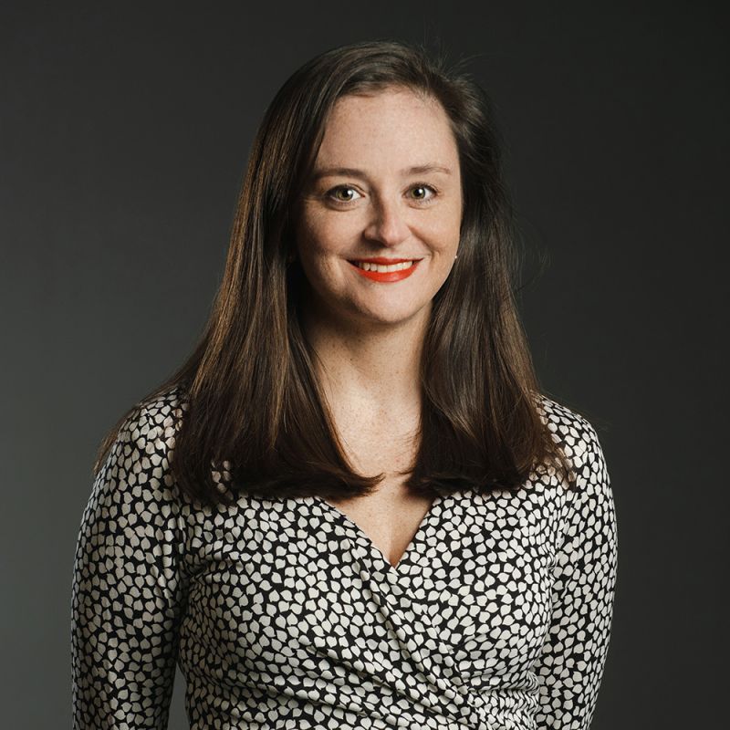 Color photograph of a light skinned woman with long brown hair wearing a black and white patterned dress and smiling at the camera