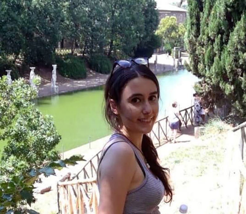 Color photograph of the head and shoulders of a light skinned woman with brown hair standing on a hill above a reflecting pool; she turns toward the camera and smiles