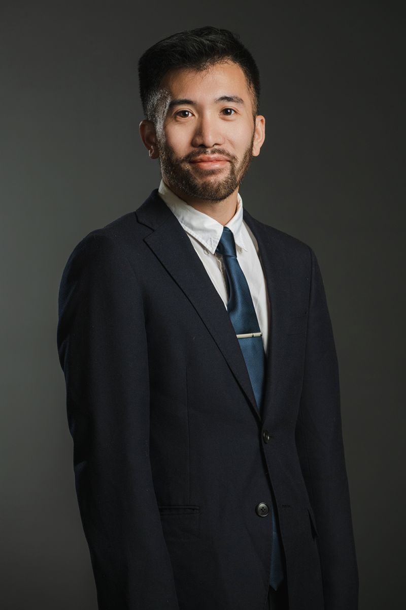 Color photograph of an Asian man with dark hair wearing a dark suit and tie as he looks at the camera