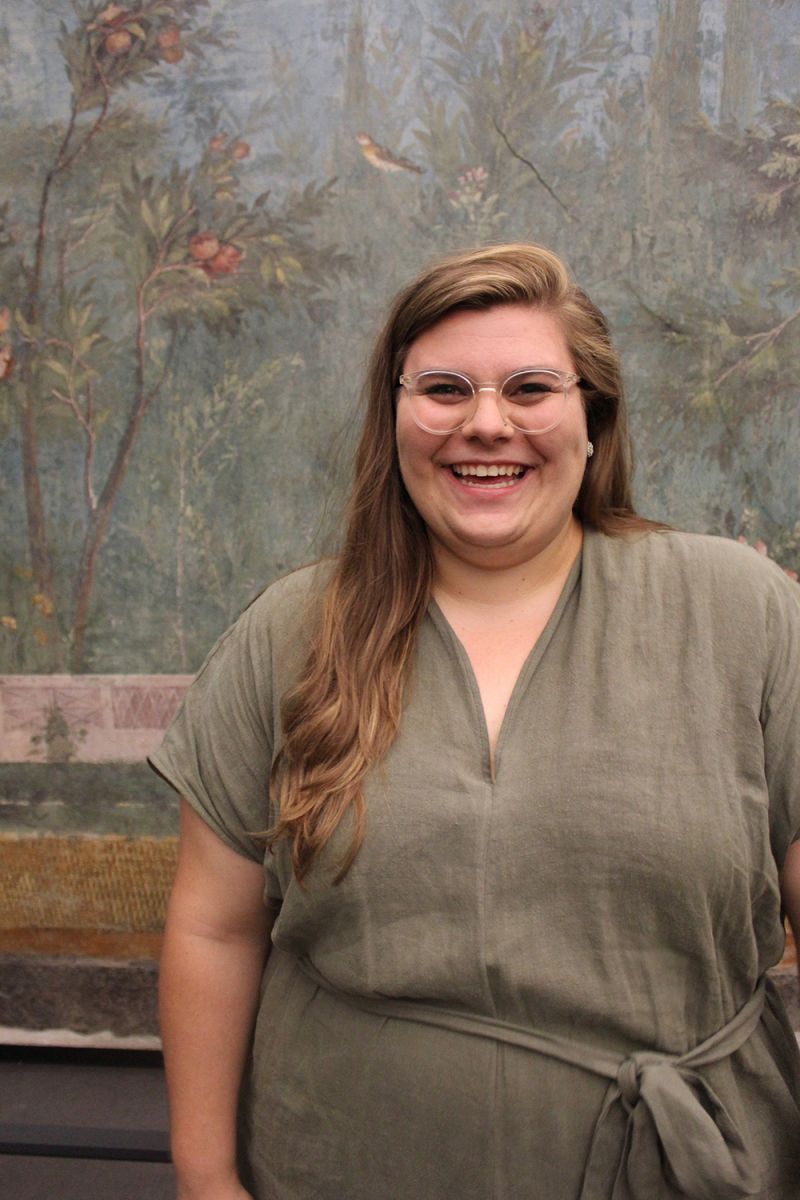 Color photo of a woman wearing a beige jumpsuit and standing in front of an ancient Roman mural