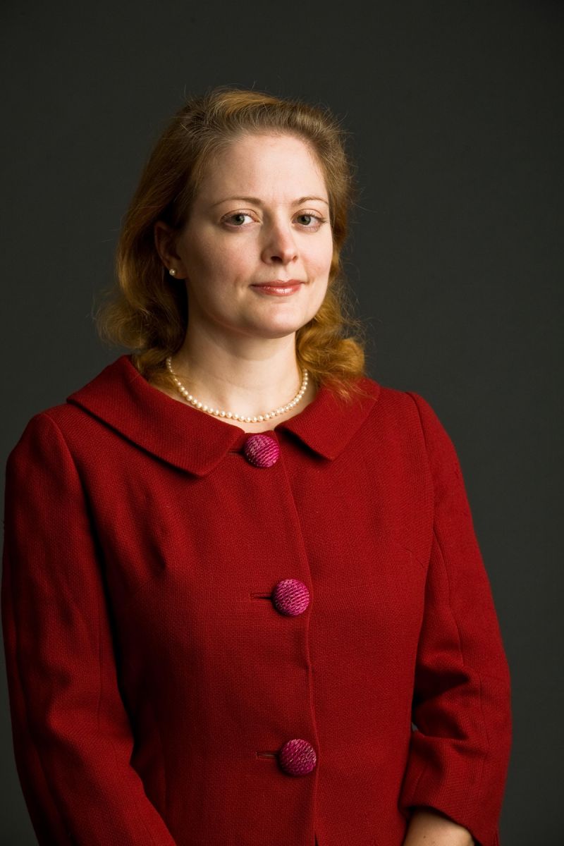 Color photograph of the head and torso of a light skinned woman in a photographer's studio wearing a red coat and smiling slightly at the camera