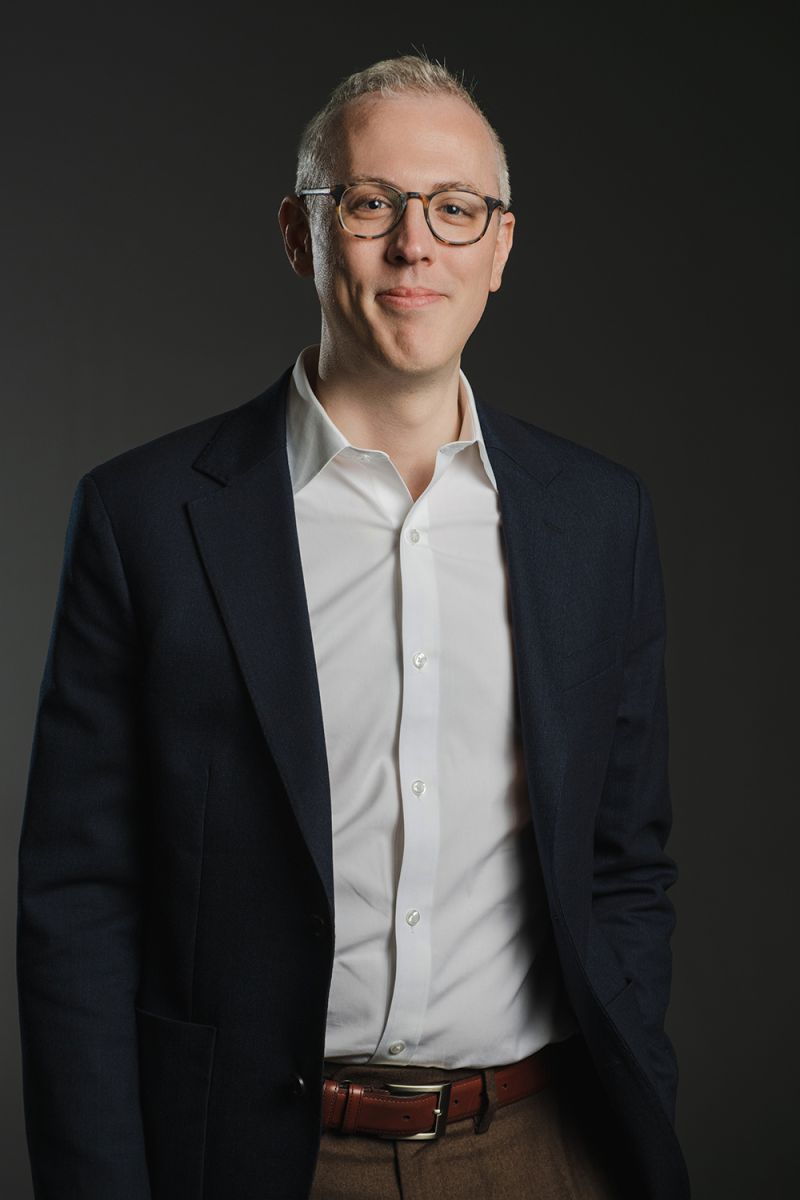 Color photograph of a light skinned man wearing glasses, a dark blazer, and a white shirt; he smiles at the camera
