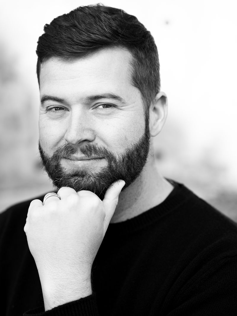 Black and white photograph of the head and chest of a light skinned bearded man; he smiles faintly at the camera and rests his chin on his hand