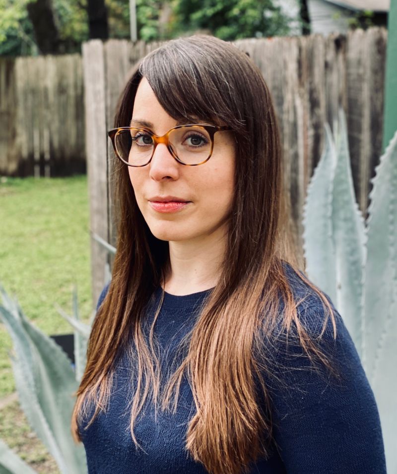 Color photographic portrait of Christy Q. Schirmer with glasses and a blue top, with a large aloe plant and a wooden fence behind her