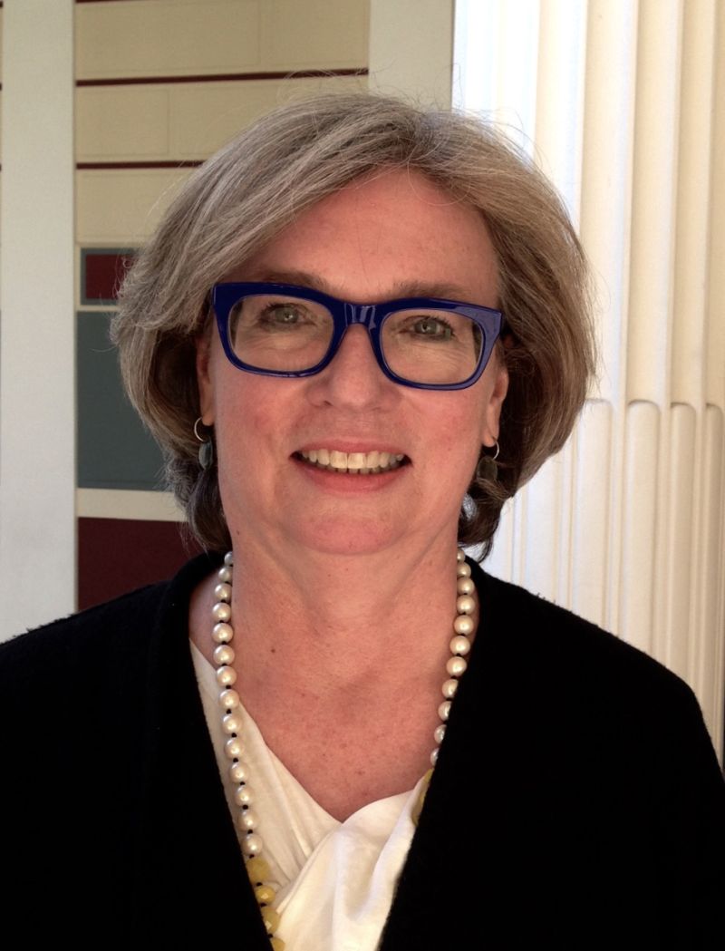 Color headshot of Claire Lyons,  a light skinned woman wearing a dark sweater, a pearl necklace, and blue eyeglasses, looking directly at the camera with a slight smile