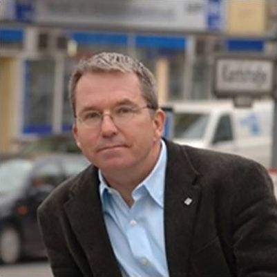 Color photograph of the head and shoulders of a light skinned man wearing a dark blazer over a blue shirt; he is standing on a city sidewalk with cars and buildings behind him