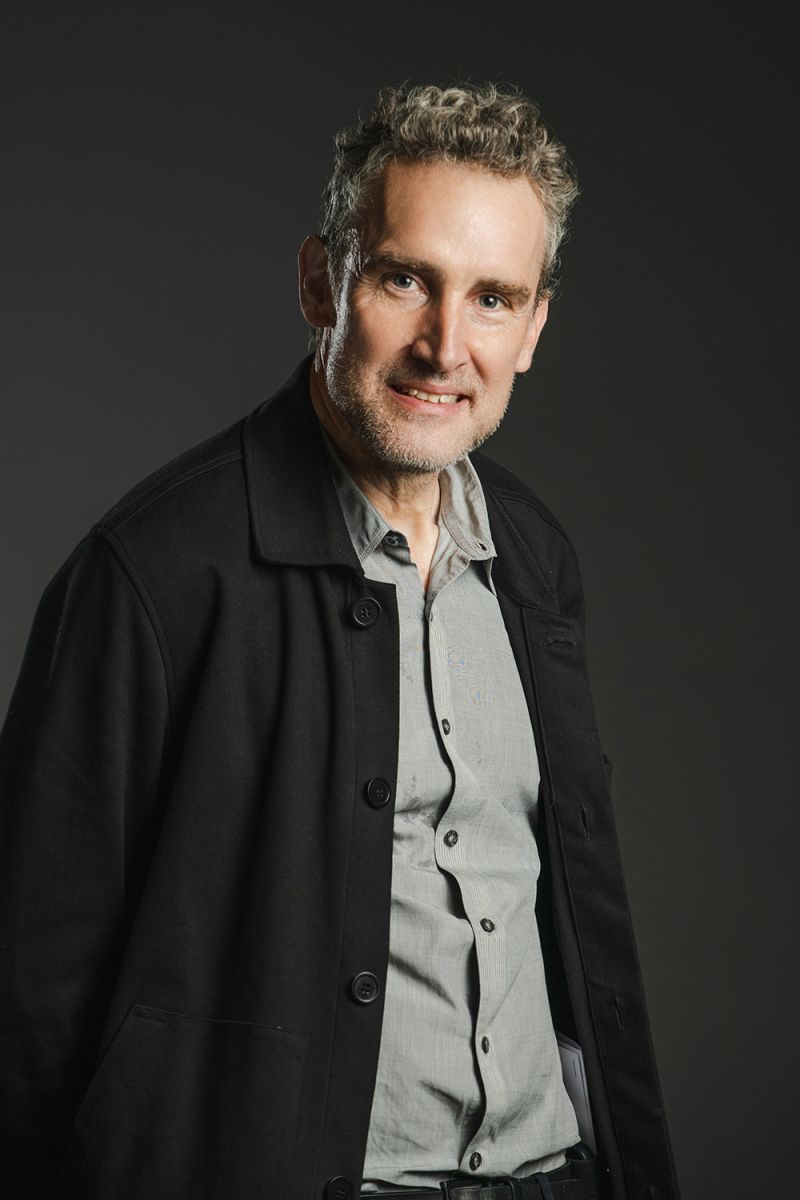 Color photograph of a light skinned man wearing a dark jacket over a button up shirt