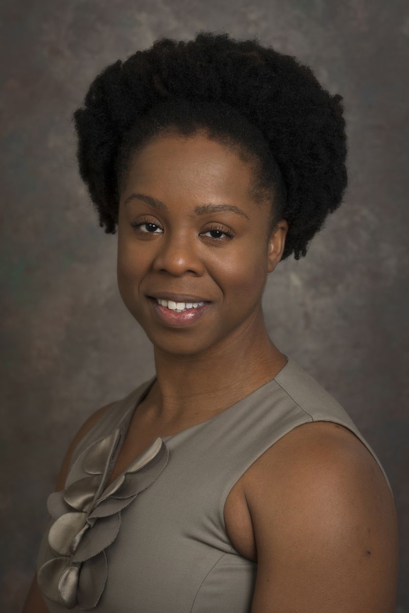 Color photograph of the head and shoulders of a dark skinned woman with a dark afro wearing a sleeveless dress; she smiles while looking directly at the camera