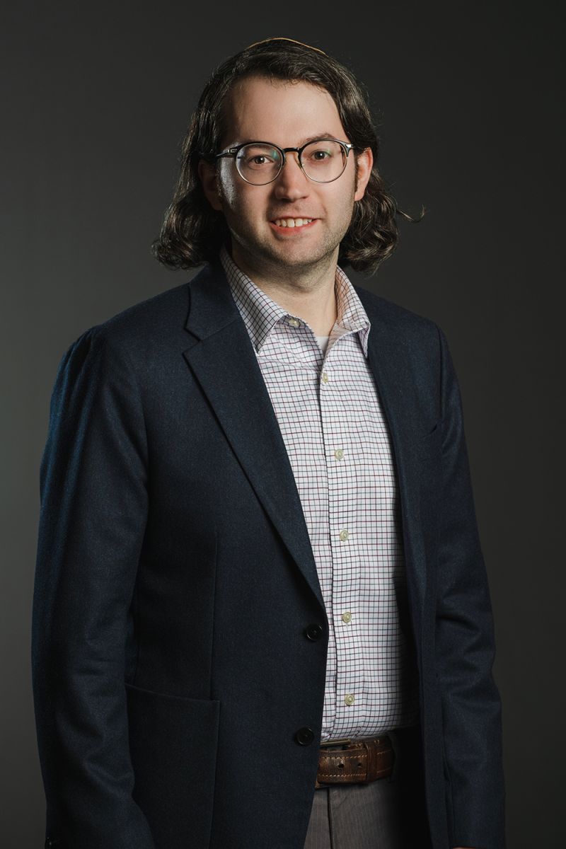 Color photograph of a light skinned man wearing glasses and a dark blazer over a plaid shirt; he looks at the camera confidently