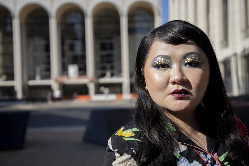 Color photograph of an Asian woman wearing colorful makeup and glitter on her face as well as a floral print top; she stares at the camera in a serious manner. Behind her are the tall arches of the opera building at Lincoln Center in New York