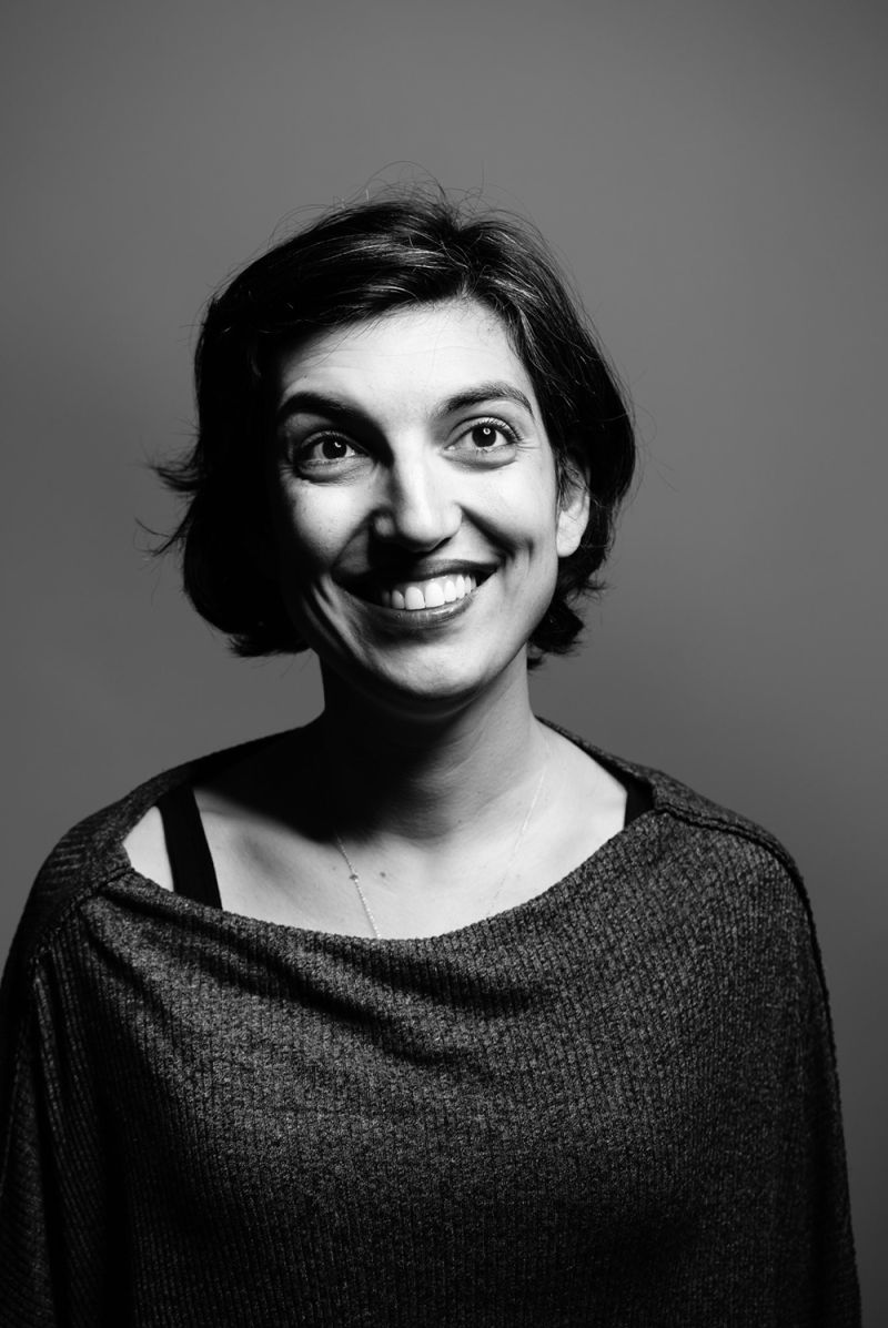 Black and white photograph of a medium skinned woman with dark hair sitting in front of a studio backdrop; she looks upward and smiles