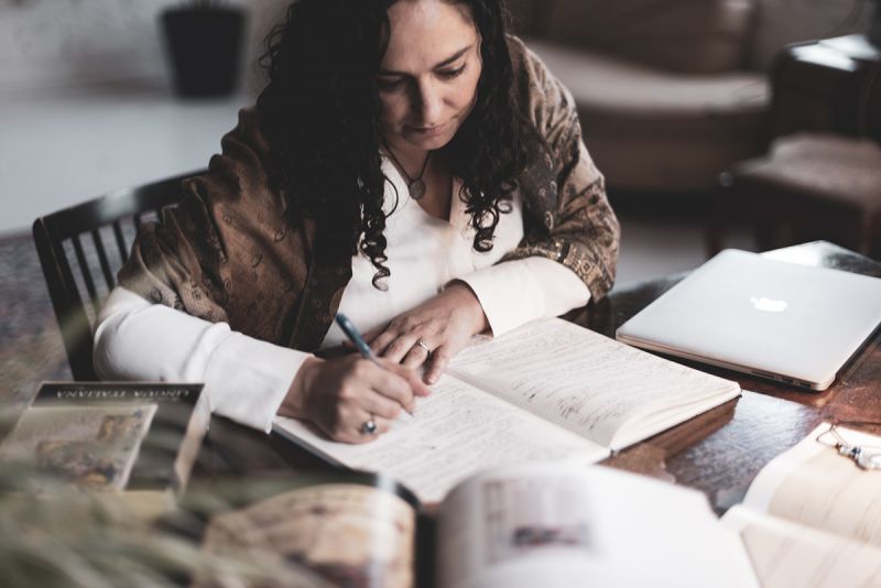 Color photograph of a light skinned woman with long brown hair sitting at a desk covered with books and papers; she holds a pen in her hand and is writing