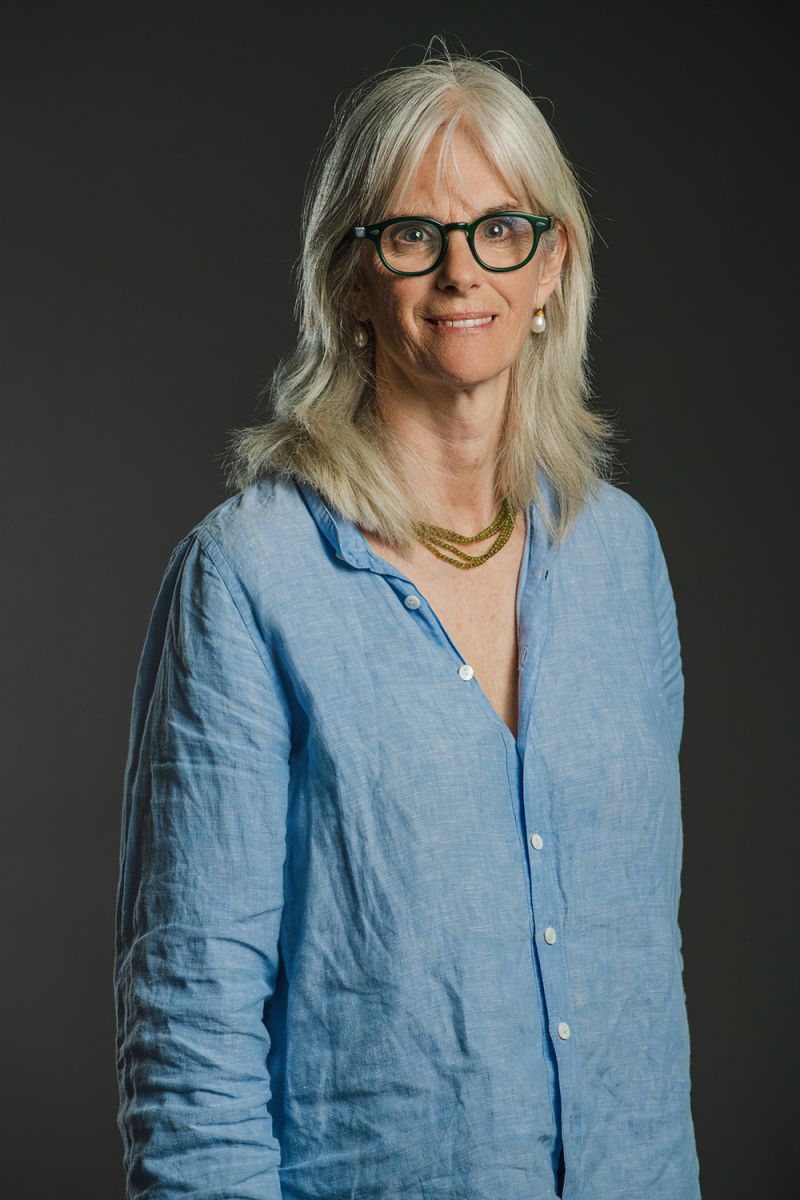 Color photograph of a light skinned woman with gray hair wearing glasses and a loose fitting blue top