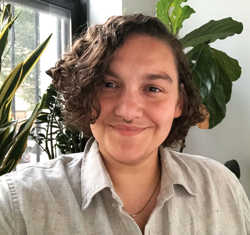 Color photograph of the head and shoulders of a light brown skinned person with dark hair smiling and looking just off center; they wear a tan linen shirt and are situated in an indoor room with green potted plants and a window 