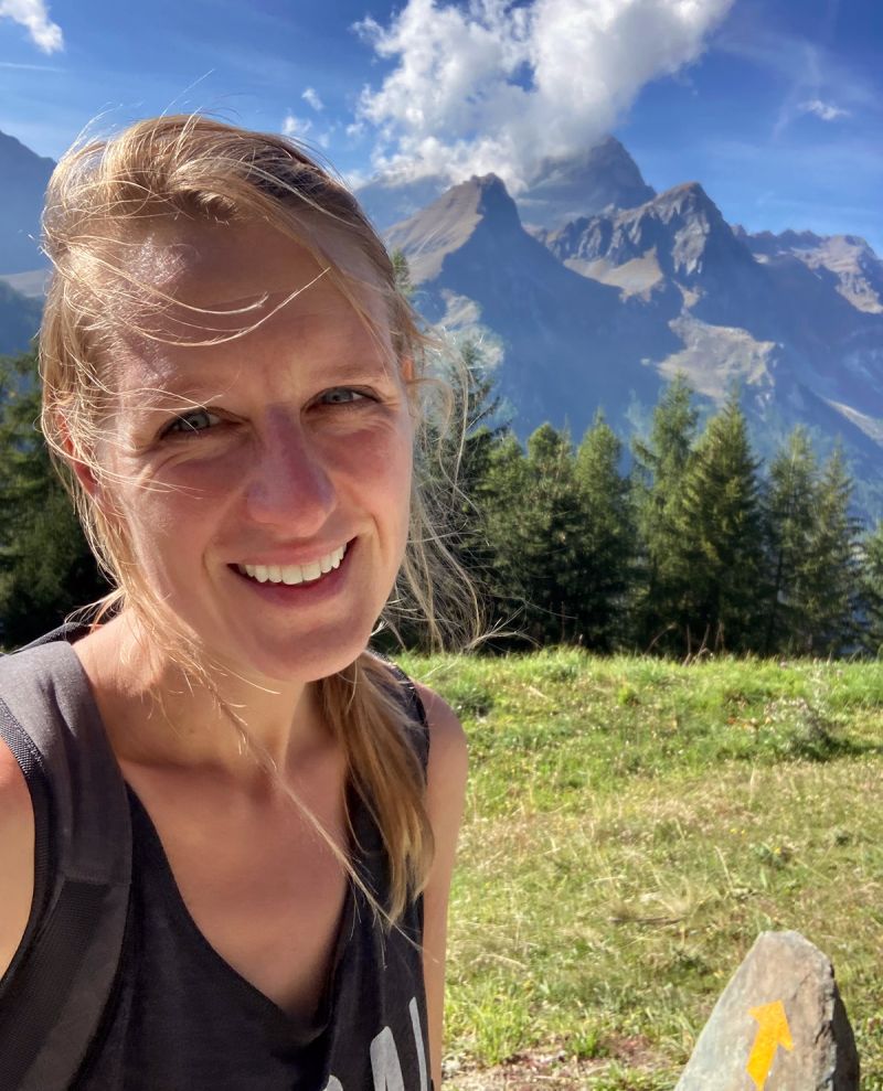 Color photograph of the head and torso of a light skinned woman with long blond hair standing in an outdoor location with a mountain in the background; she smiles directly at the camera