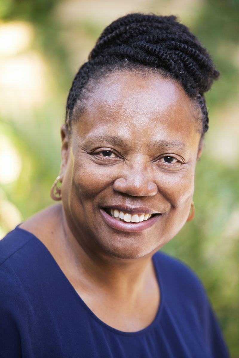Color photograph of a dark skinned woman located in an outdoor location wearing a blue shirt and smiling at the camera