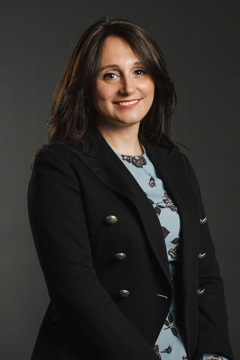 Color photograph of a light skinned woman with brown hair wearing a blue blazer with big buttons over a blue botanical print shirt; she smiles directly at the camera