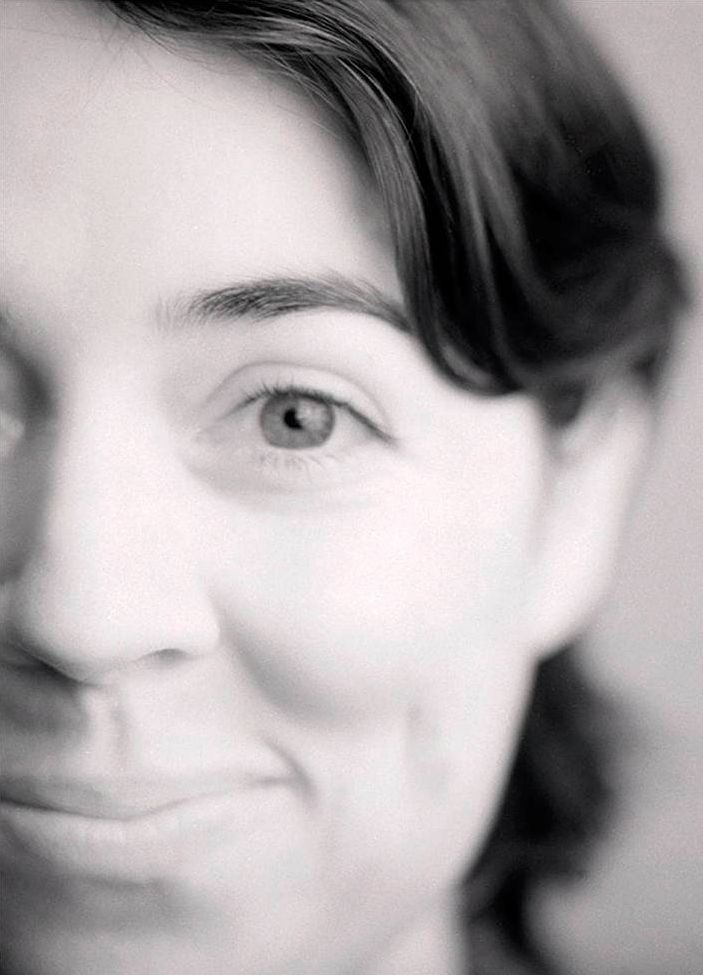 Black and white photograph of the face of a light skinned woman smiling with a closed mouth; the image is cropped just past her nose on the left side