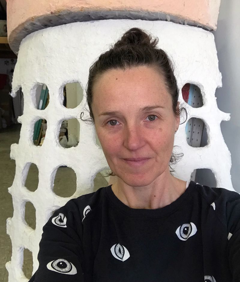 Color photograph of the head and shoulders of a light skinned woman with her dark hair tied back in a bun; she wears a dark shirt with prints of eyes on it and stands in front of a ceramic sculpture painted white