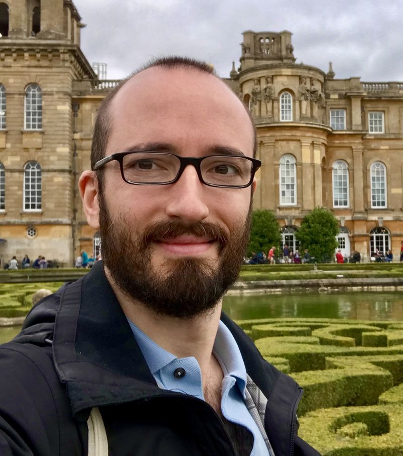 Color photograph of the head and shoulders of Eugenio Refini standing in an outdoor courtyard with a church or university building behind him