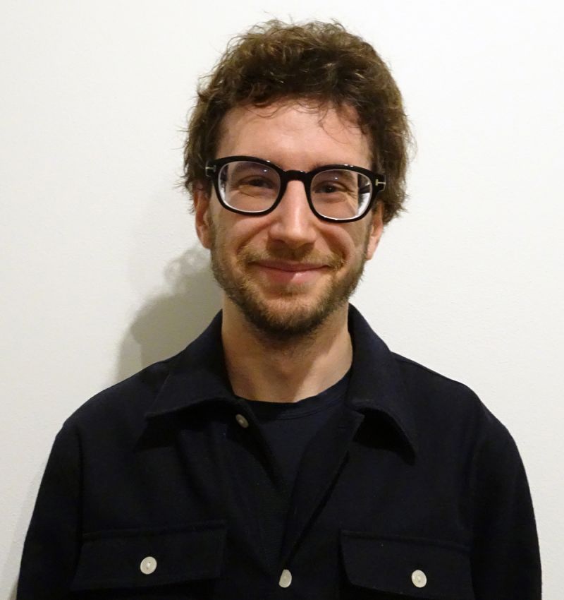 Color photo of a light skinned man with dark hair and glasses smiling at the camera