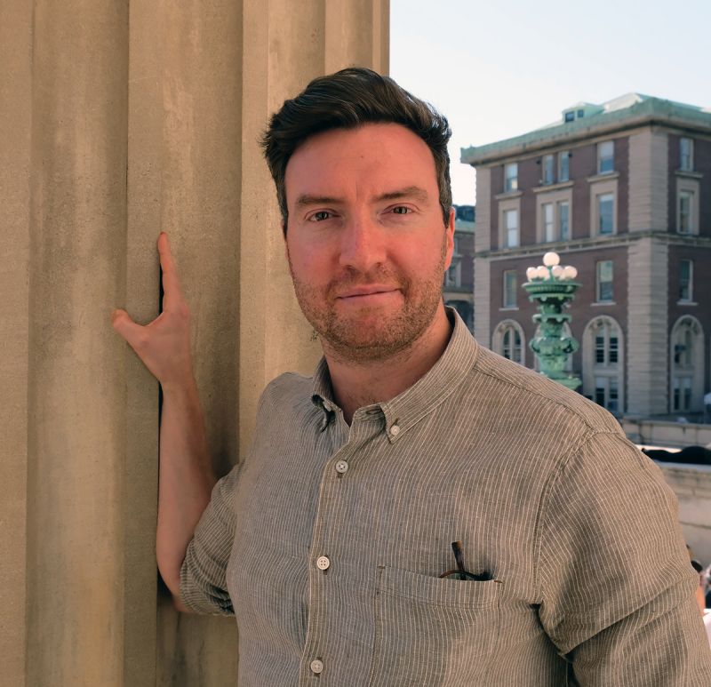Color photograph of a light skinned man with brown hair and a gray button-up shirt standing next to a fluted stone column, on which his hand rests; he looks directly at the camera