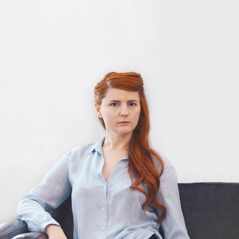 Color photograph of a light skinned woman with long red hair wearing a button-up shirt and looking at the camera