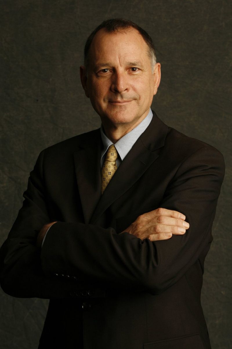 Color photograph of the head and torso of a light skinned man wearing a dark suit and tie and smiling slightly at the camera