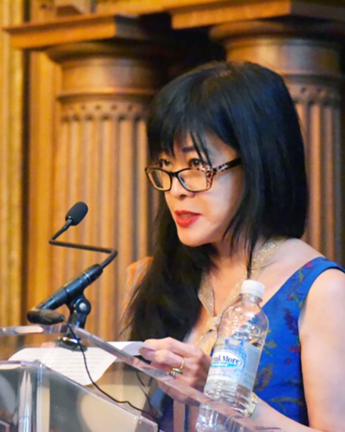 Color photo of an Asian woman wearing a blue dress and glasses and standing at a podium with a microphone