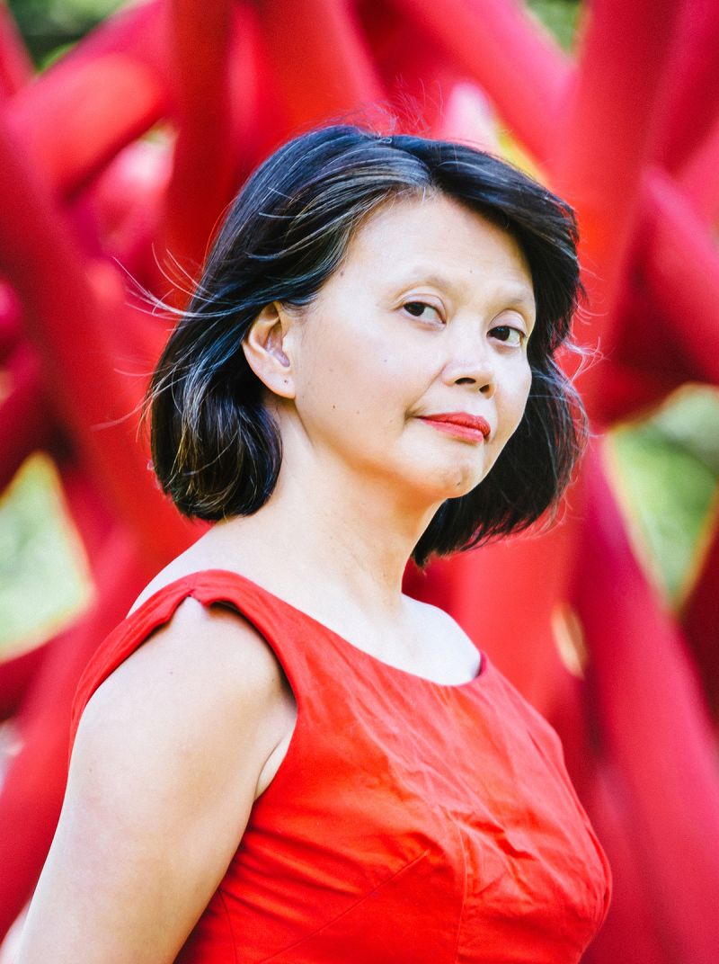 Color photograph of the head and torso of a light skinned Filipino woman with dark hair, wearing a wrinkled red dress and looking at the camera; she stands in front of what looks like a very large but blurred foliage