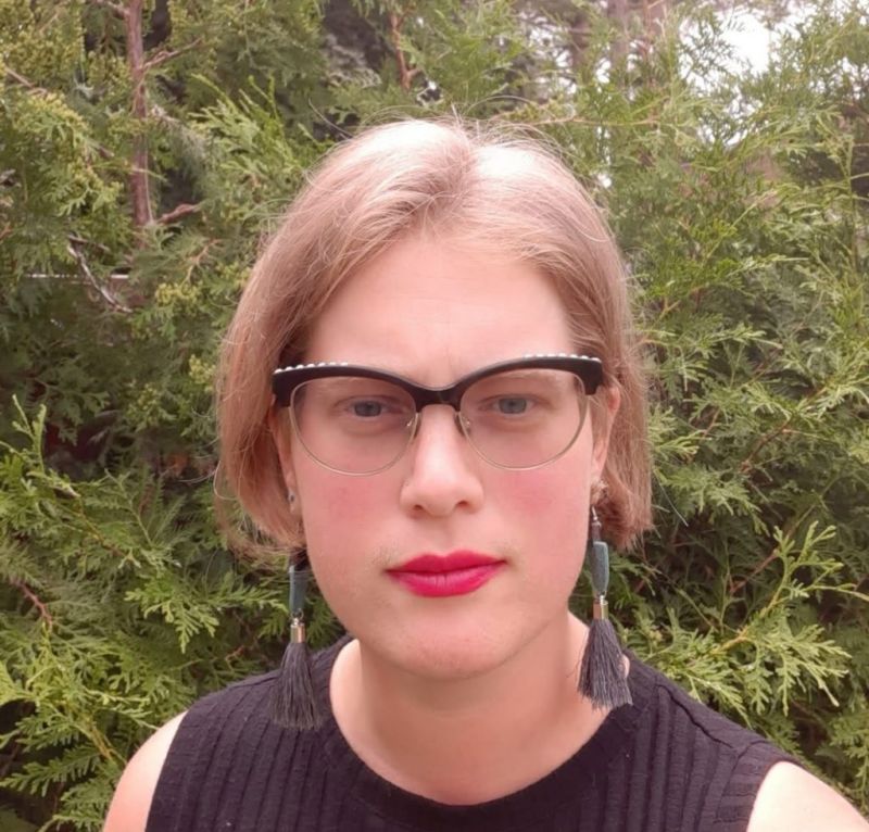Color photograph of the head and shoulders of Gloria Bell, standing outside with a green foliage behind her