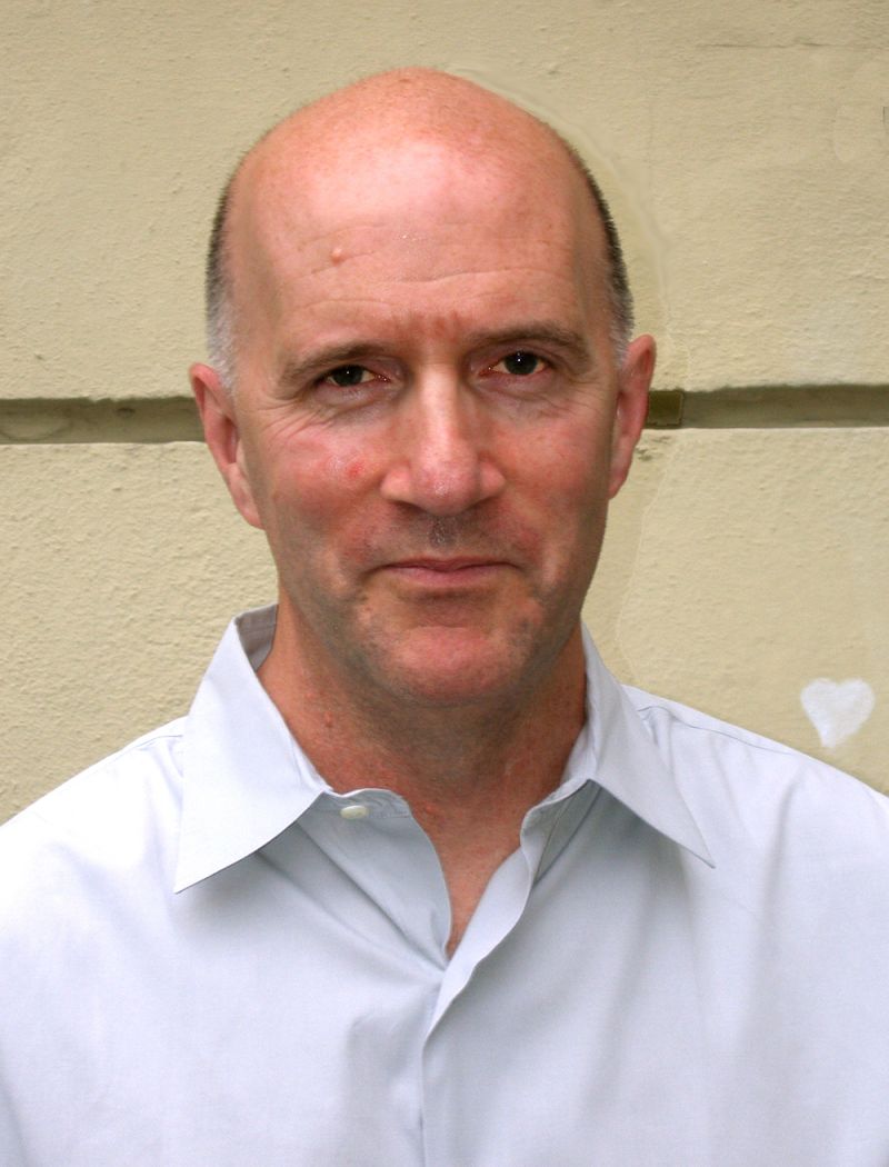 Color photograph of the head and torso of a light skinned bald man looking at the camera with a serious expression