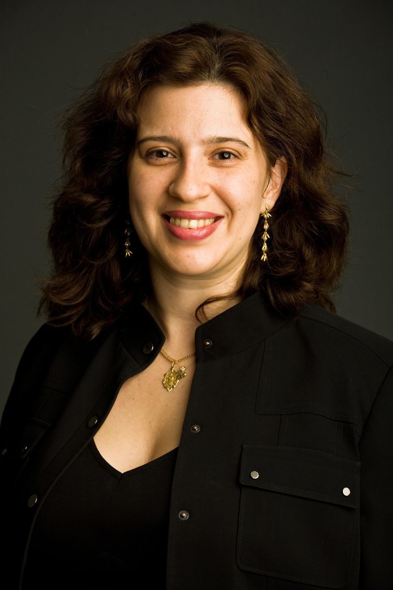 Color photograph of the head and torso of a light skinned woman in a photographer's studio wearing a black top and blazer and smiling at the camera