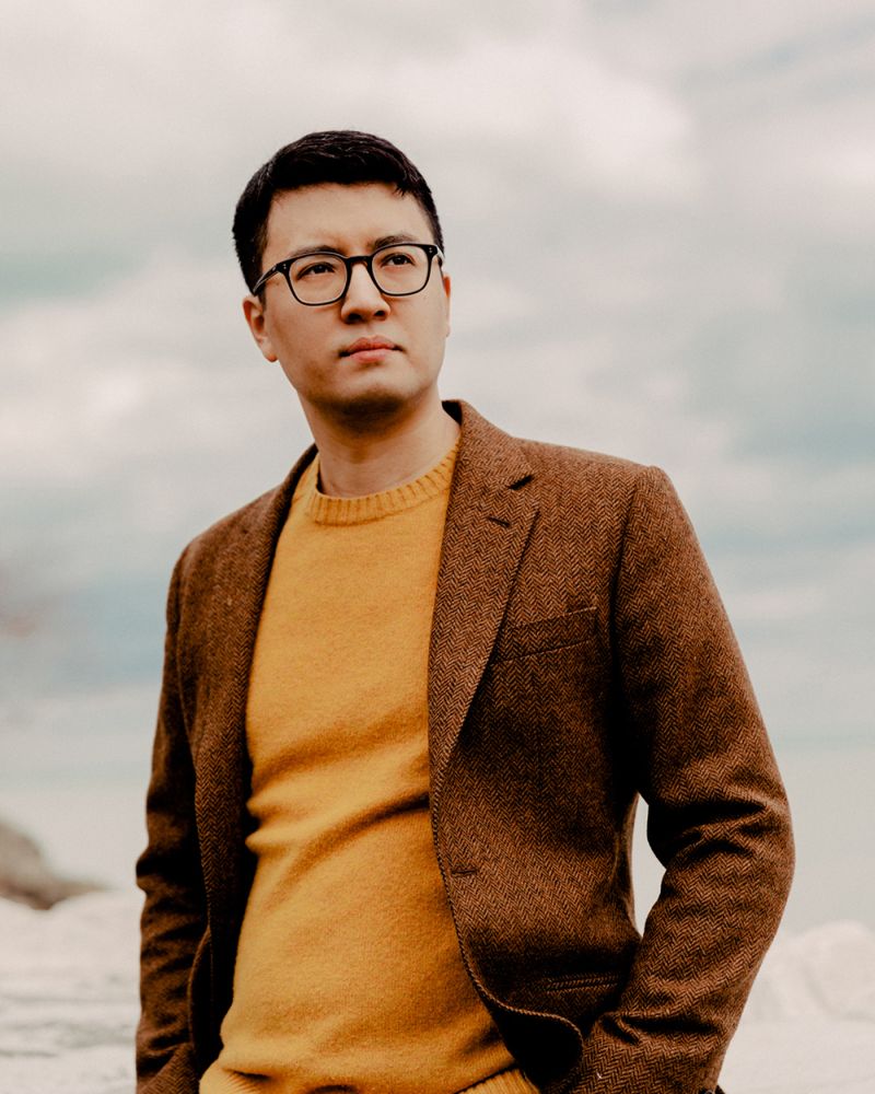 Color photograph of Igor Santos standing outside, wearing an orange sweater and burgundy blazer